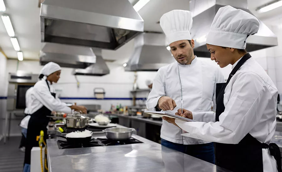 chefs in a professional kitchen setting, appearing to be discussing or reviewing something on a digital tablet.