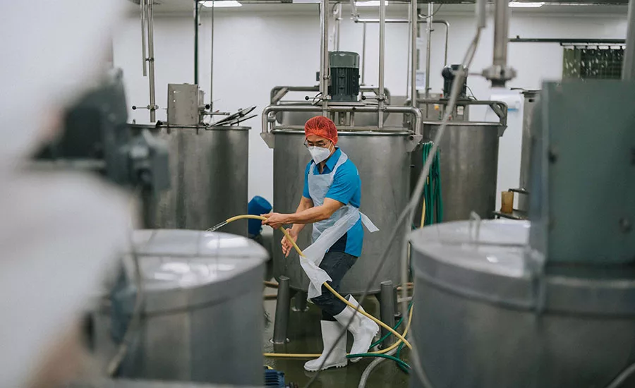 a worker cleaning the floor around large stainless steel processing vats in an industrial food production facility, potentially a winery or cheesery