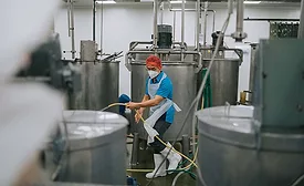 a worker cleaning the floor around large stainless steel processing vats in an industrial food production facility, potentially a winery or cheesery