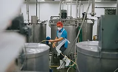 a worker cleaning the floor around large stainless steel processing vats in an industrial food production facility, potentially a winery or cheesery