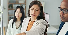 This image shows a woman in a white blazer actively collaborating with a team in an office setting.