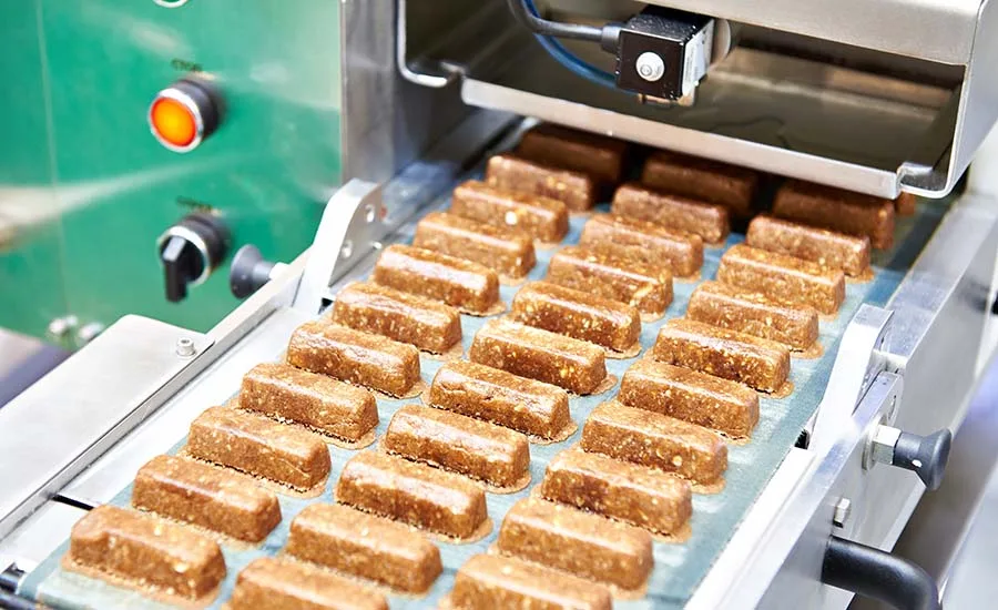 a stainless-steel industrial conveyor belt carrying rows of uniform, brownish rectangular bars