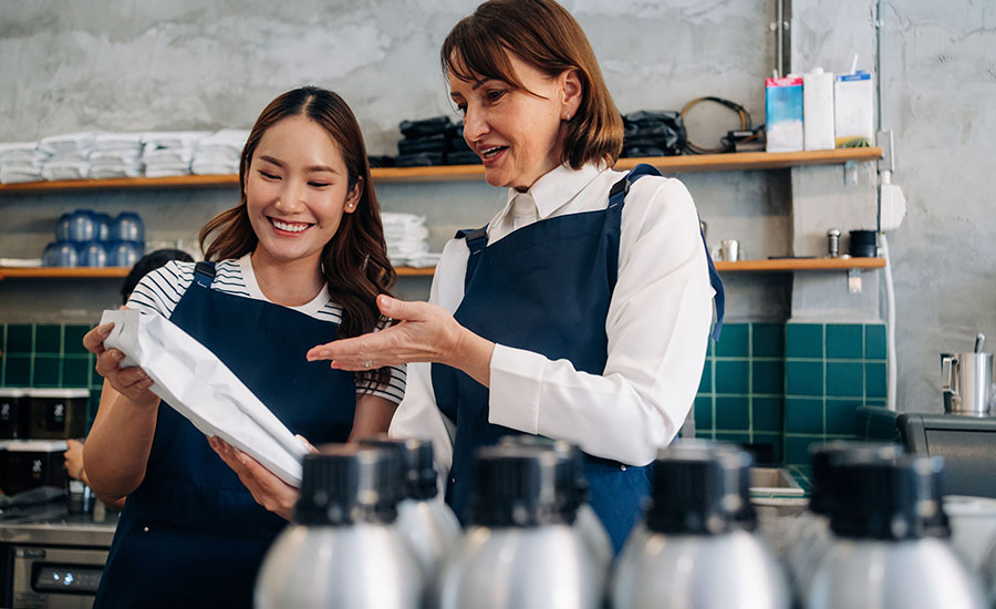 two cafe workers wearing navy blue aprons while reviewing inventory or supplies behind a counter