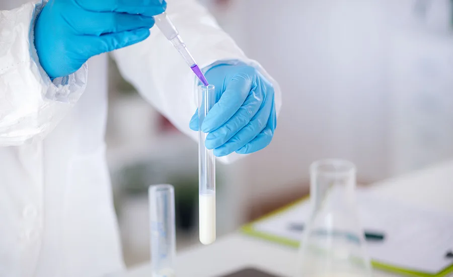 a person wearing a white lab coat and blue gloves using a pipette to dispense a purple liquid into a glass test tube containing a white, milky substance a person wearing a white lab coat and blue gloves using a pipette to dispense a purple liquid into a glass test tube containing a white, milky substance