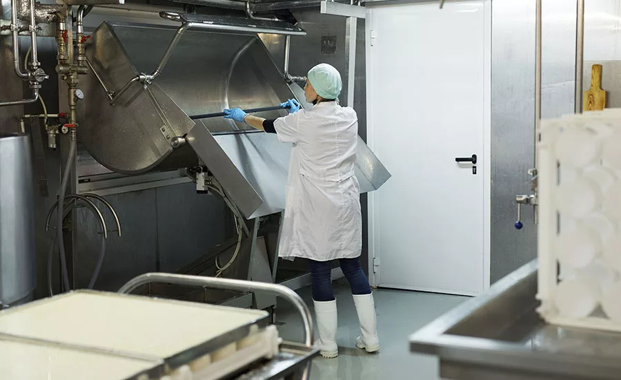 The image shows a worker cleaning a industrial cheesemaking curdling tank within a dairy factory. 