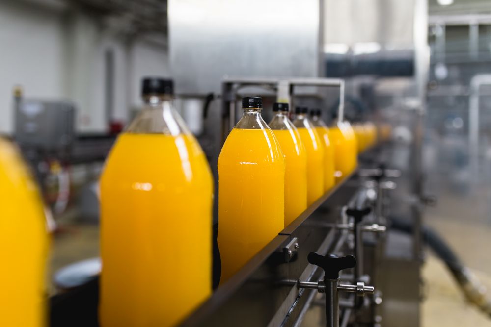 The image shows plastic bottles filled with orange juice moving along an automated production and conveyor line in a bottling factory