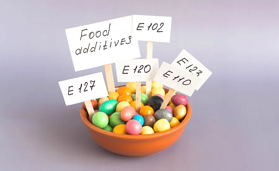 a small bowl filled with colorful candy-like granules and paper signs on wooden sticks labeled with European food additive code numbers like E 102, E 110, E 120, E 123, and E 127 a small bowl filled with colorful candy-like granules and paper signs on wooden sticks labeled with European food additive code numbers like E 102, E 110, E 120, E 123, and E 127
