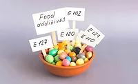 a small bowl filled with colorful candy-like granules and paper signs on wooden sticks labeled with European food additive code numbers like E 102, E 110, E 120, E 123, and E 127 a small bowl filled with colorful candy-like granules and paper signs on wooden sticks labeled with European food additive code numbers like E 102, E 110, E 120, E 123, and E 127
