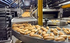 The image depicts a high-volume industrial baking factory line where freshly baked bread rolls are moving along a conveyor system. 