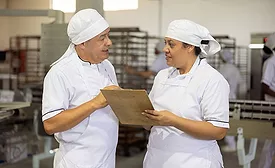 This image shows two food production professionals in white uniforms reviewing safety or quality documentation on a clipboard within a manufacturing facility