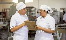 This image shows two food production professionals in white uniforms reviewing safety or quality documentation on a clipboard within a manufacturing facility