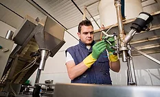 This image depicts a professional mechanic working on a piece of stainless steel processing machinery in a factory setting. This image depicts a professional mechanic working on a piece of stainless steel processing machinery in a factory setting.