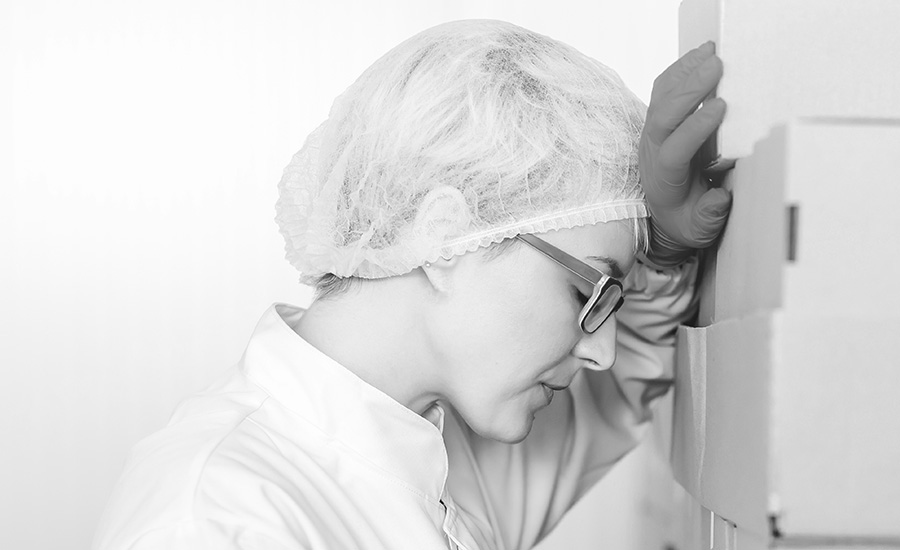 a person wearing a protective hairnet, glasses, and a white lab coat or gown, appearing stressed, tired, or overwhelmed while leaning against a surface