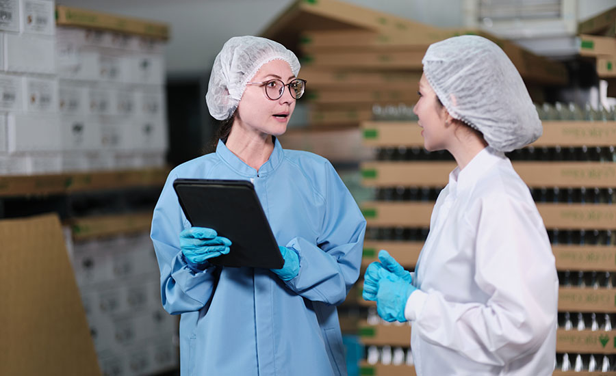 two professionals wearing hairnets, protective coats, and blue gloves inside a manufacturing or storage facility, with one holding a tablet