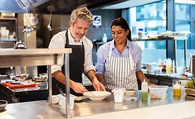 two individuals wearing aprons working together at a stainless-steel counter inside a commercial kitchen