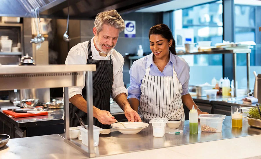 two individuals wearing aprons working together at a stainless-steel counter inside a commercial kitchen two individuals wearing aprons working together at a stainless-steel counter inside a commercial kitchen