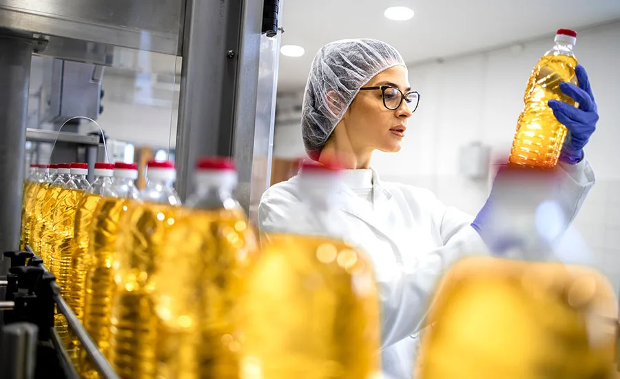 a worker in a hairnet and lab coat inspecting a production line of bottled cooking oil
