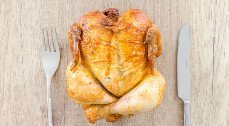whole roast chicken on wood table in between fork and knife