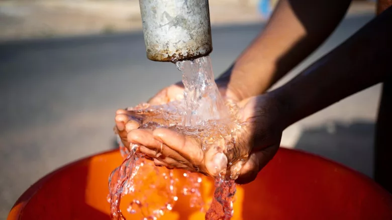person holding their hands cupped under water running from pipe outdoors into bucket