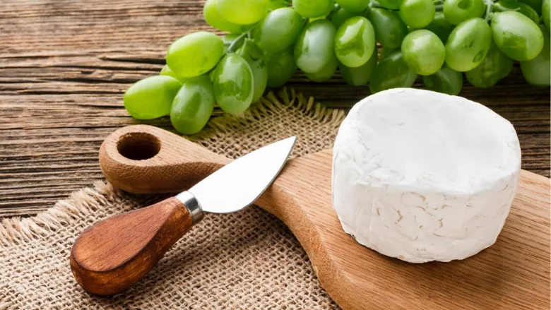 small wheel of goat cheese on charcuterie board next to cheese knife and green grapes
