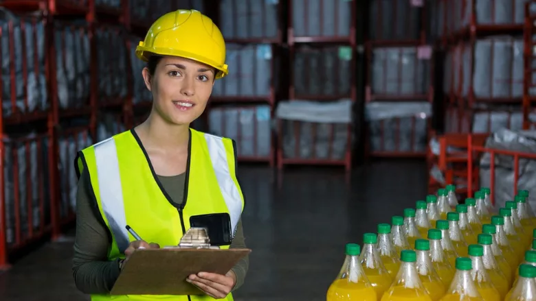 woman in hard hat and vest holding clipboard in warehouse next to pallet of bottled beverages
