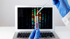 gloved hands holding pipette and tube in front of laptop with genetic sequencing data in background