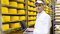 man working in a cheese warehouse smiling at camera holding laptop