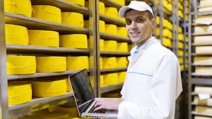 man using laptop in cheese warehouse_usertrmk via Freepik.png man working in a cheese warehouse smiling at camera holding laptop