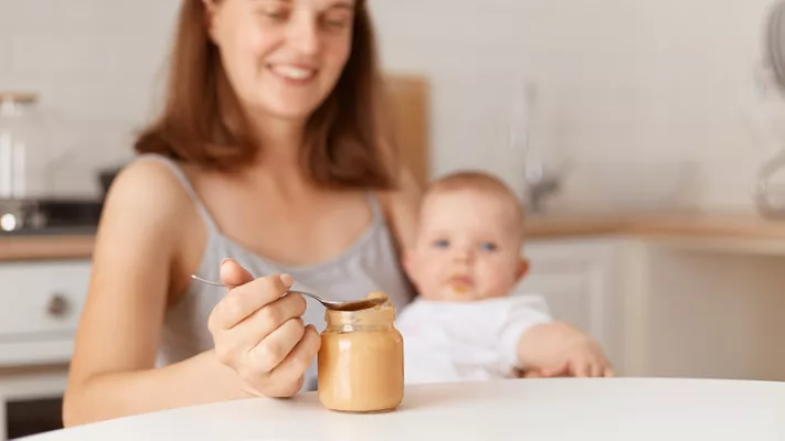 mom baby and jar of baby food_user18526052 via Magnific.png jar of baby food on table in foreground with mom holding baby in background