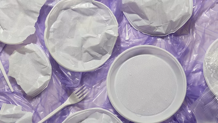 plastic utensils and plates on crumpled plastic film against purple background