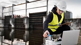 scientist in yellow vest and hard hat kneeling down next to water in wastewater treatment plant collecting samples with laptop and clipboard