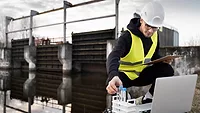 scientist in yellow vest and hard hat kneeling down next to water in wastewater treatment plant collecting samples with laptop and clipboard
