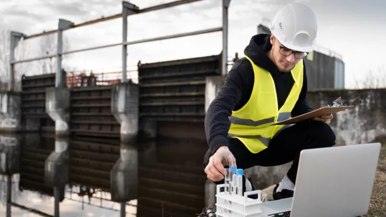 scientist in yellow vest and hard hat kneeling down next to water in wastewater treatment plant collecting samples with laptop and clipboard