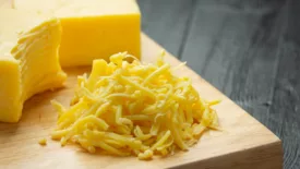 half shredded block of cheddar cheese and pile of cheese shreds on wooden cutting board