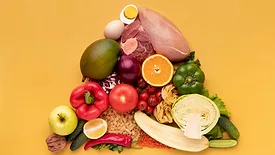 a variety of whole foods including vegetables, meats, nuts, bread, and pasta arranged in a pyramid shape laid against a yellow backdrop