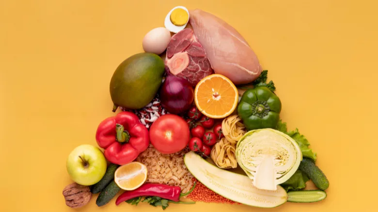 a variety of whole foods including vegetables, meats, nuts, bread, and pasta arranged in a pyramid shape laid against a yellow backdrop