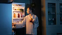 a woman in pajamas looking at the fridge grabbing snacks in a dark kitchen