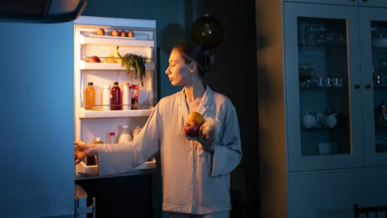 a woman in pajamas looking at the fridge grabbing snacks in a dark kitchen