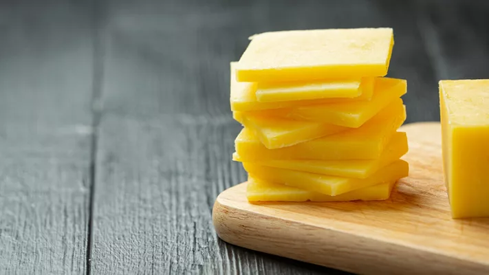 slices off a block of cheddar cheese on a wooden cutting board