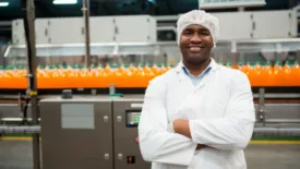 smiling employee in bottling factory looking at camera with arms crossed in front of production line